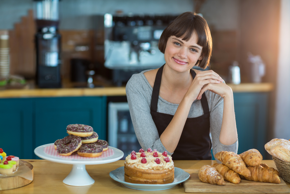 Portrait of waitress sitting at counter with sweet food on table in cafx92xA9