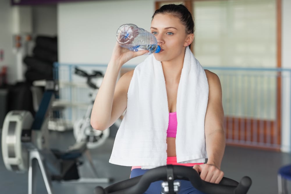 Tired young woman drinking water while working out at exercise bike in gym